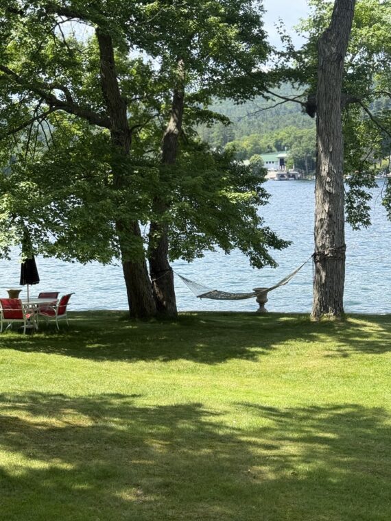 A hammock is tied between two trees on a grassy lawn beside a lake, with a set of red and white chairs and a table nearby. Lush green trees frame the view, and buildings are visible across the water.