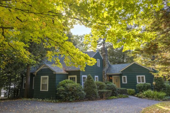 A green cottage-style house sits among lush trees with sunlight filtering through bright green leaves, casting dappled light on the driveway and surrounding shrubs.