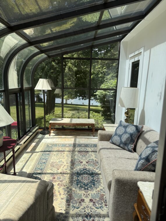 A sunlit conservatory features large glass windows and ceiling, a patterned rug, a beige sofa with blue pillows, two standing lamps, a bench, and views of trees and a lake outside.