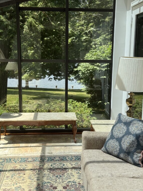 Sunlight streams into a cozy living room with a sofa, patterned pillow, table lamp, and a marble coffee table. Large windows offer a view of a green lawn, trees, and water outside.