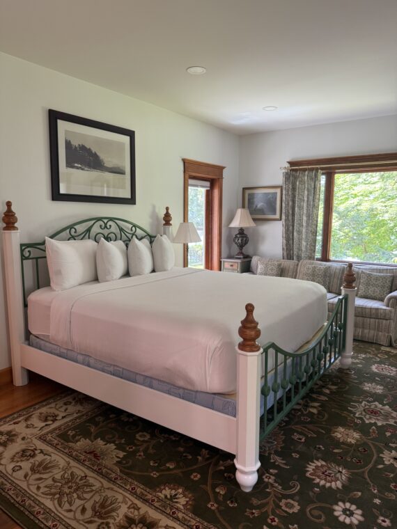 A bright bedroom with a large bed featuring a green and white metal frame, white linens, and several pillows. There is a sofa, large window with trees outside, lamps, and a framed landscape photo on the wall.