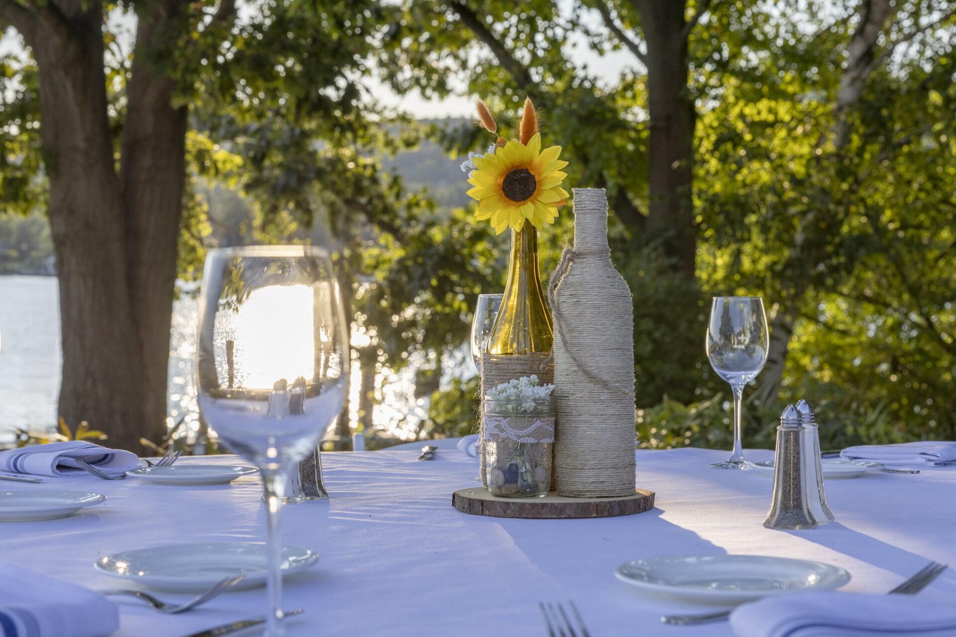 A beautifully set outdoor dining table with a white tablecloth, decorated with a centerpiece featuring sunflowers in bottles. Wine glasses and small plates are arranged around the table. The background shows a scenic view of trees and a lake.