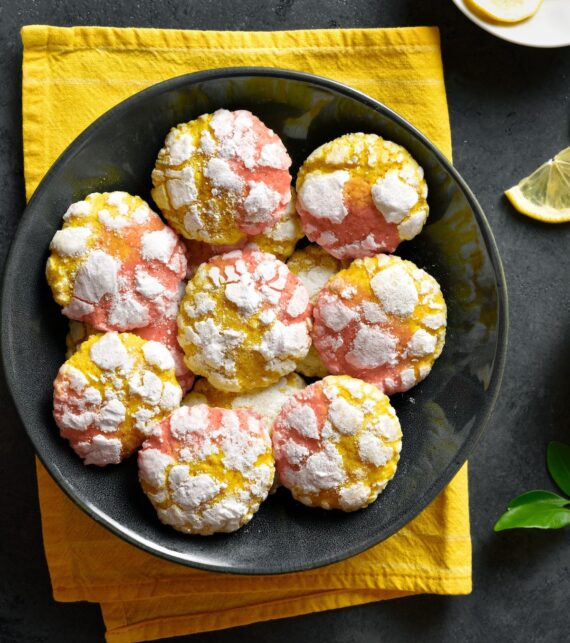 A black plate of yellow and pink crinkle cookies topped with powdered sugar sits on a yellow napkin. Lemon slices and green leaves are nearby on a dark surface.