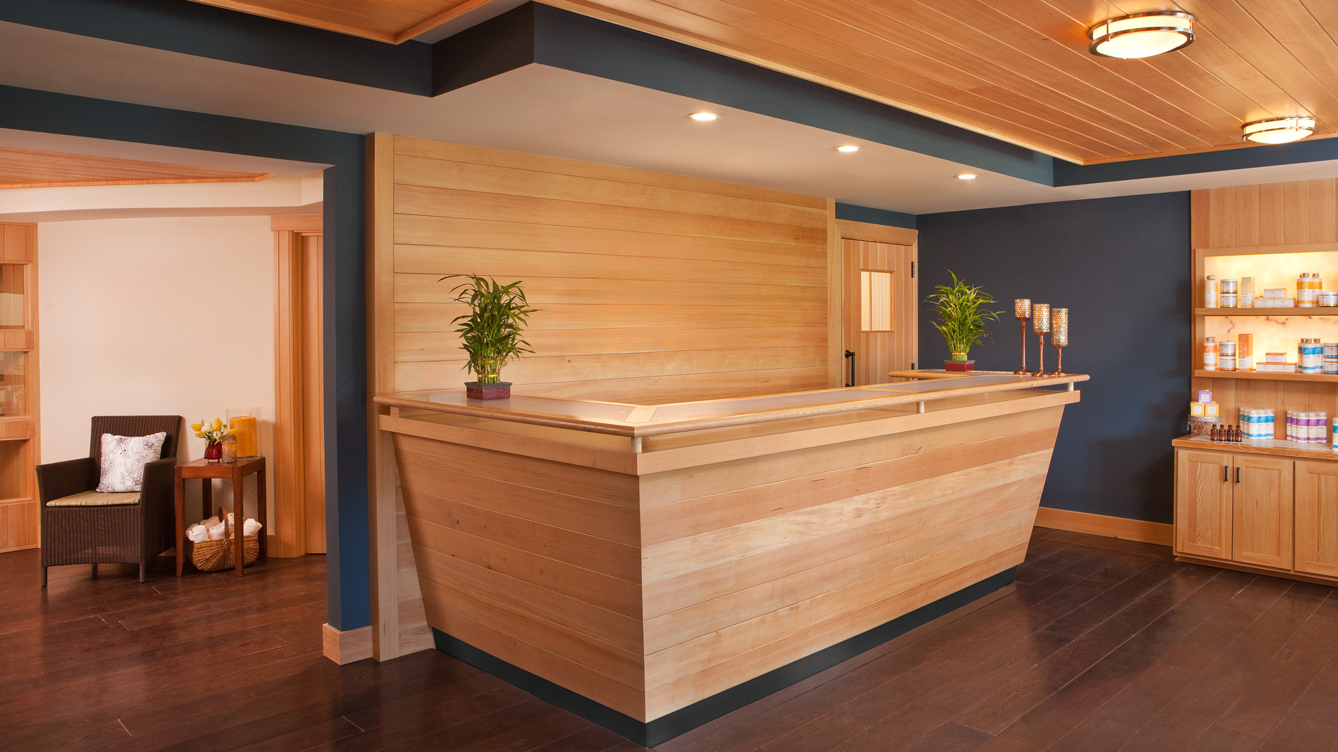 A modern spa reception area with a wooden front desk, two plants, and a blue accent wall. Shelving displays various spa products. Warm lighting and wooden ceilings create a welcoming ambiance. A seating area with a chair and small table is visible.