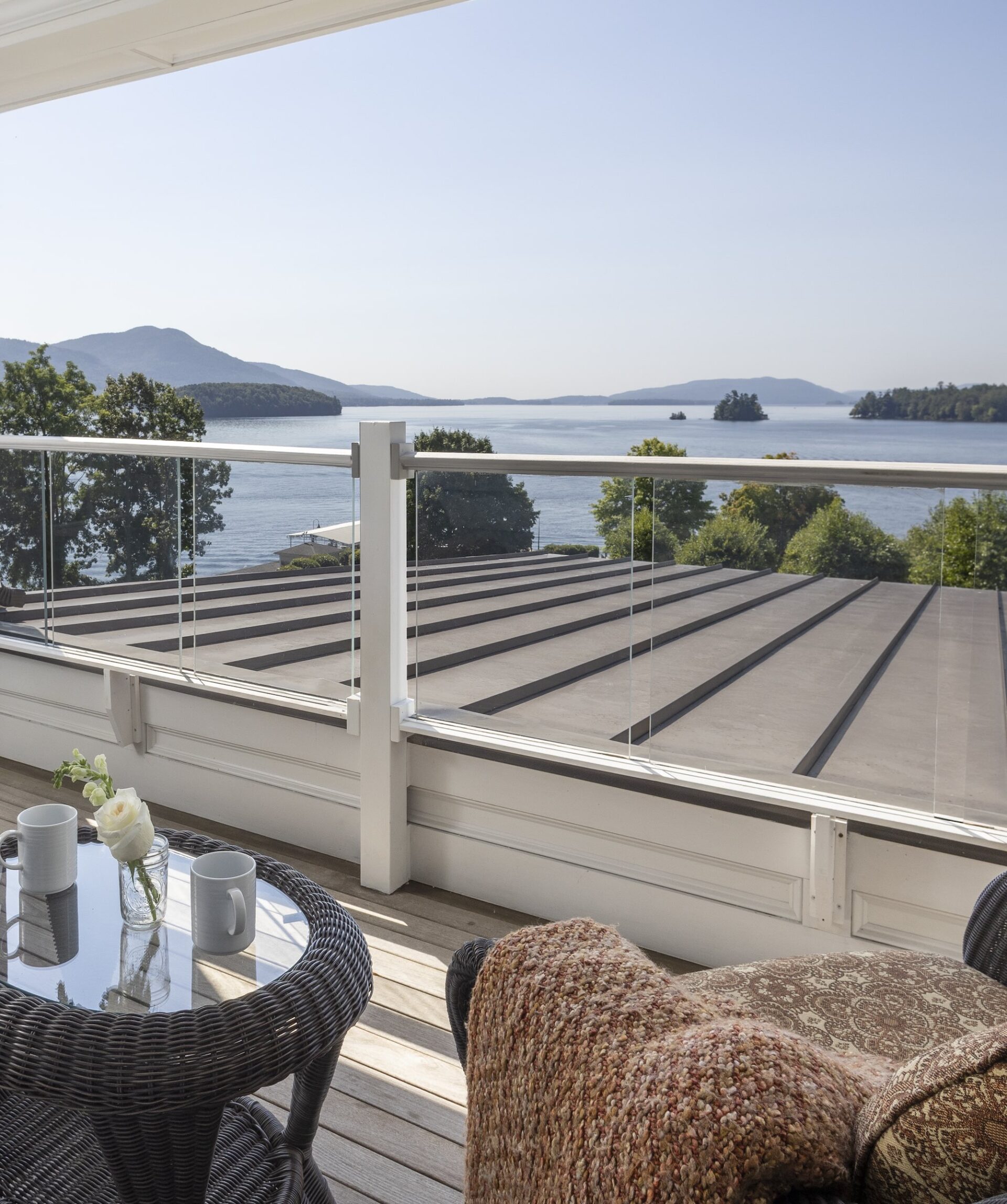 A balcony view overlooking a serene lake with distant mountains under a clear sky. The foreground features a wicker table and chairs, with white mugs and a vase with a rose, as well as a cozy blanket draped over a chair.
