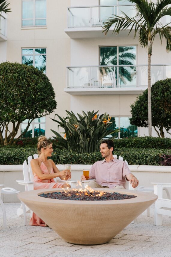 A woman and a man sit by a round outdoor fire pit, holding drinks and talking. They are surrounded by tropical plants and palm trees, with a modern building and balconies in the background.
