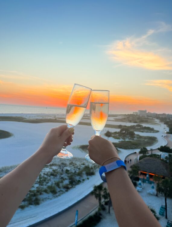 Two hands holding champagne glasses for a toast, with a beach, ocean, and colorful sunset in the background. One person wears a blue wristband.
