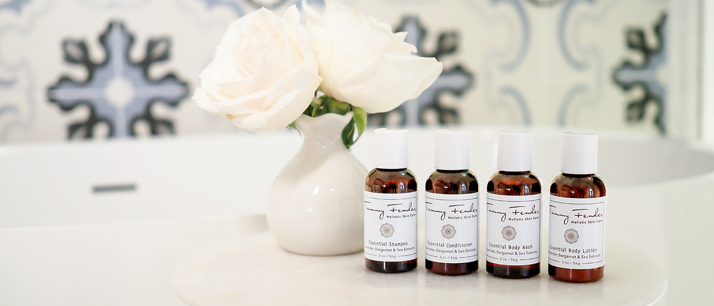 Four small amber bottles with white labels are arranged on a marble surface next to a white vase holding two white roses, with a blurred bathtub and decorative tiled wall in the background.