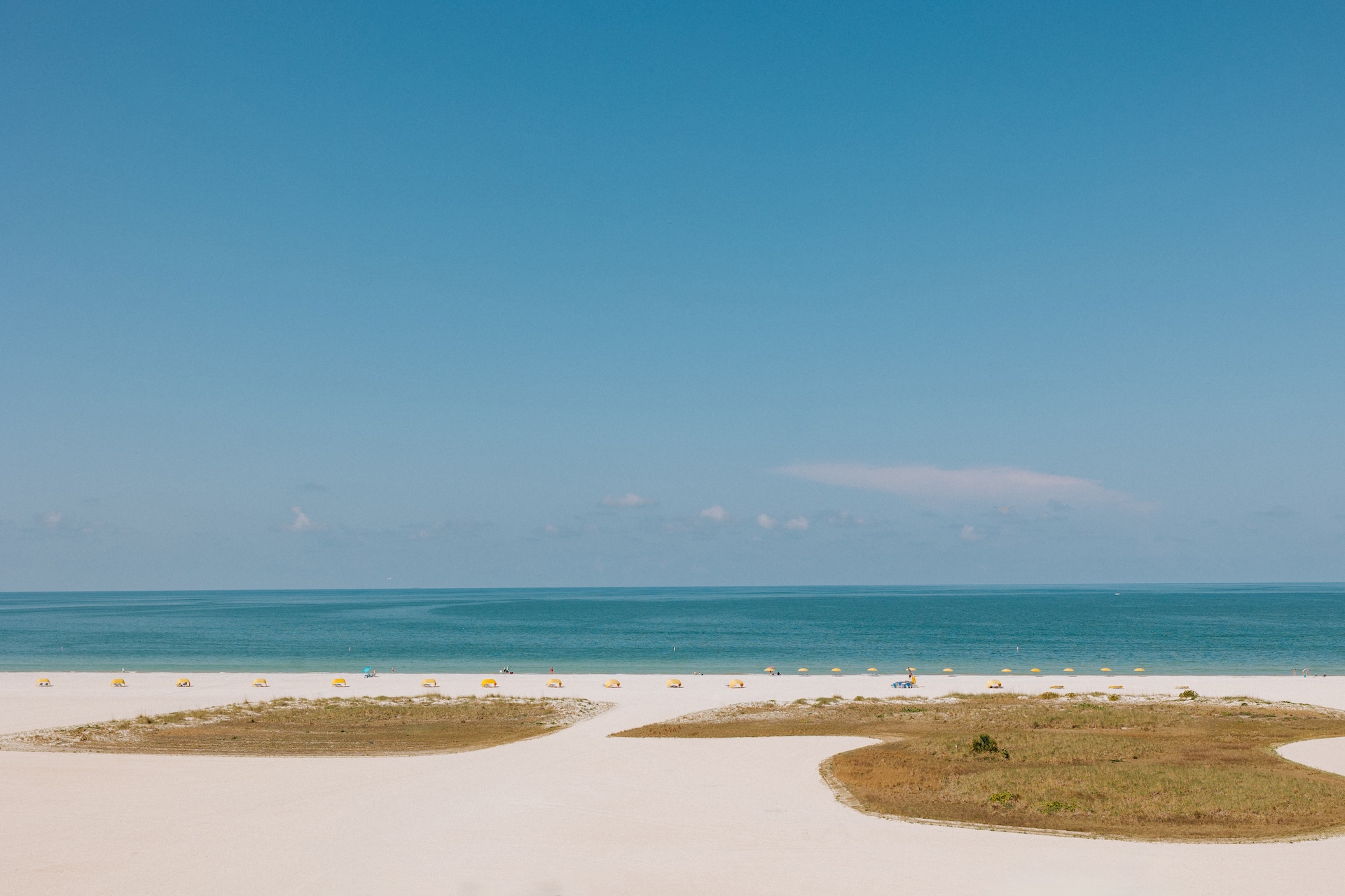 A wide sandy beach with patches of grass, yellow umbrellas lined up near the shoreline, and calm blue ocean under a clear sky.