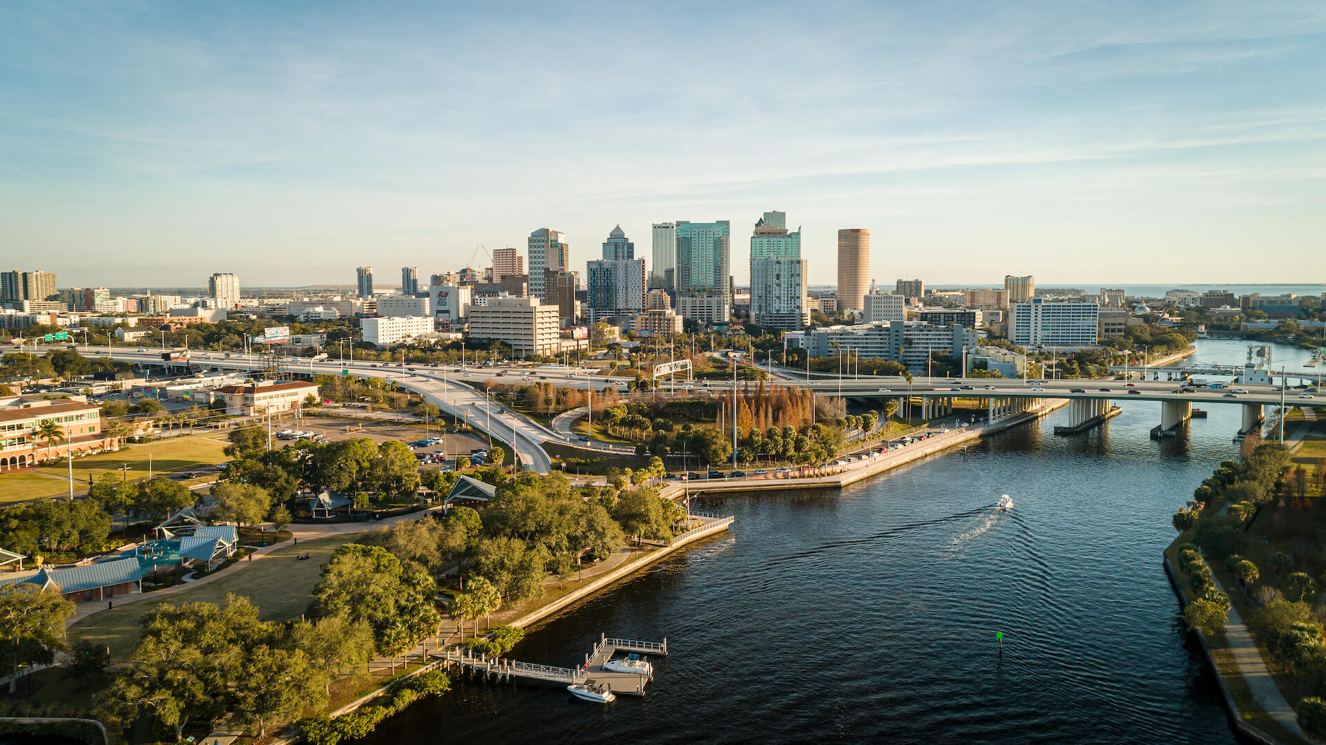 Aerial view of a city skyline with tall buildings, a river, a bridge, green spaces, and boats on the water under a clear sky.