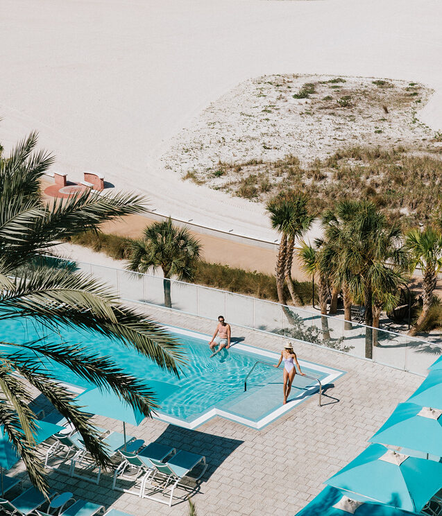 View of a beachside pool area with two people in the pool, palm trees nearby, white sand beach, clear blue ocean, and rows of yellow umbrellas in the distance under a bright sky.