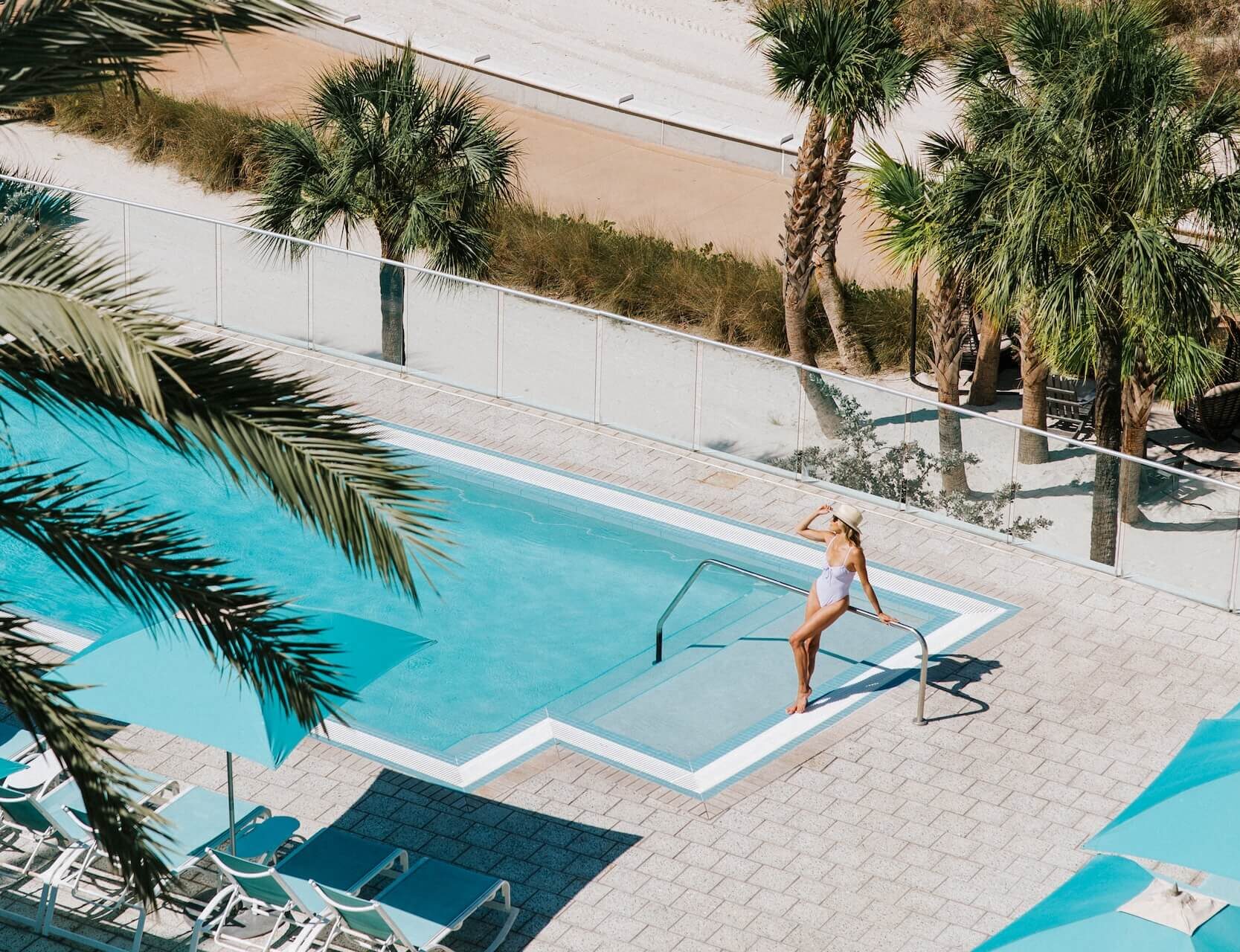 A woman in a white swimsuit stands at the edge of a swimming pool, surrounded by palm trees and blue lounge chairs, on a sunny day at a resort.