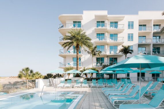 A row of light blue lounge chairs and umbrellas beside a swimming pool at a beachfront hotel.