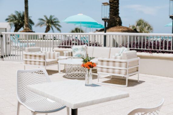 A small vase of orange flowers on a square patio table, with lounge chairs and sofas in the background on a pool deck.