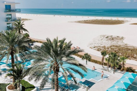 An elevated view of a swimming pool at a beachfront hotel with lounge chairs and people relaxing near the ocean.
