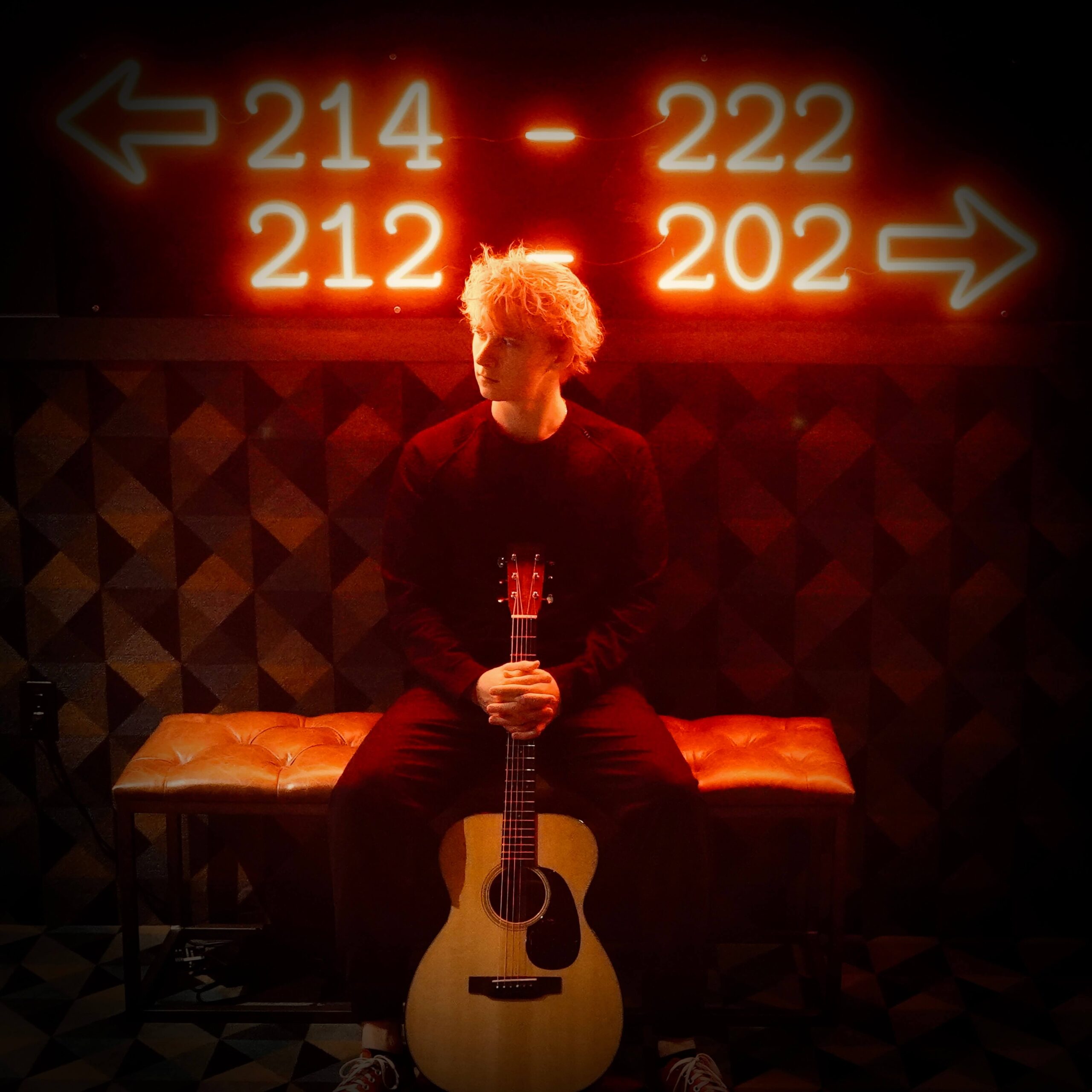 A person with light-colored hair sits on a bench holding an acoustic guitar, under bright orange neon signs with arrows and room numbers: “214 - 222” (left) and “212 - 202” (right), against a dark background.