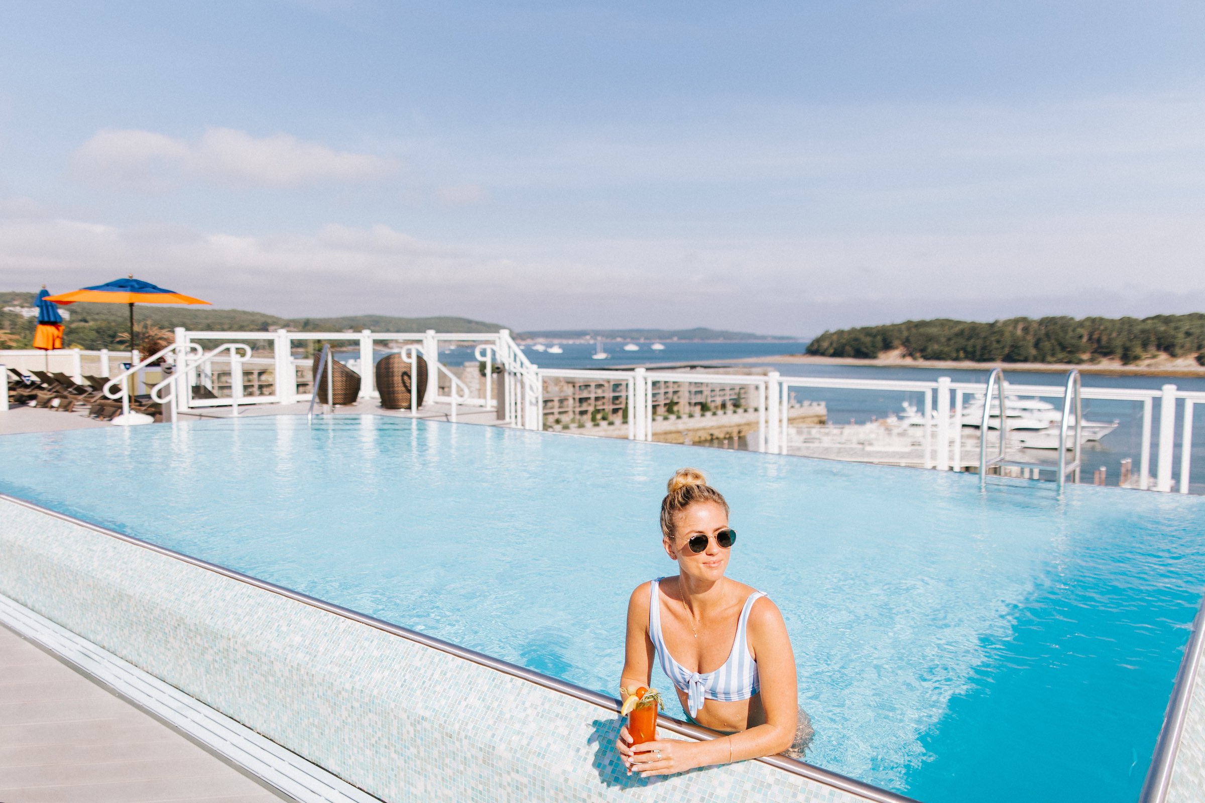 A woman in sunglasses relaxes in a swimming pool, holding a red drink. The pool overlooks a scenic view of the ocean with yachts and trees in the background. The sky is clear and sunny.