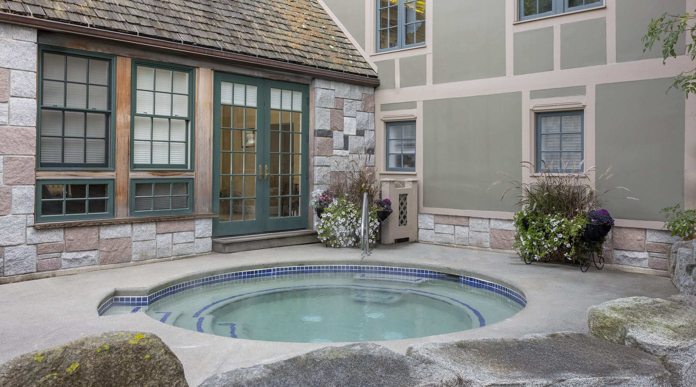 Small circular outdoor hot tub in a stone-paved courtyard, surrounded by stone and wood-paneled walls with windows. Decorative plants in pots are placed around the area.