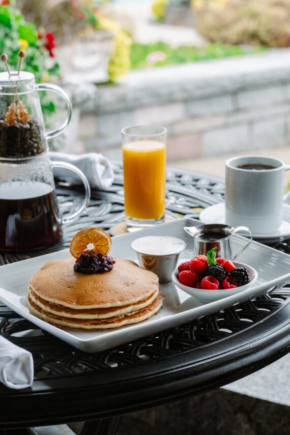 A breakfast table with a stack of pancakes topped with fruit and syrup, a small bowl of mixed berries, a cup of coffee, a glass of orange juice, and a pot of coffee on an ornate metal table outdoors.