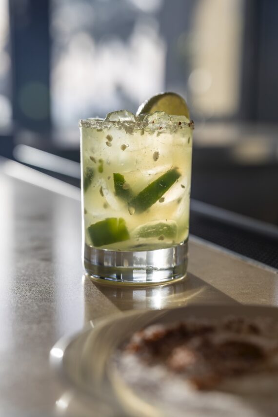 A glass of cocktail with lime slices, ice, and salt on the rim sits on a sunlit surface, with a blurred plate of spices in the foreground.