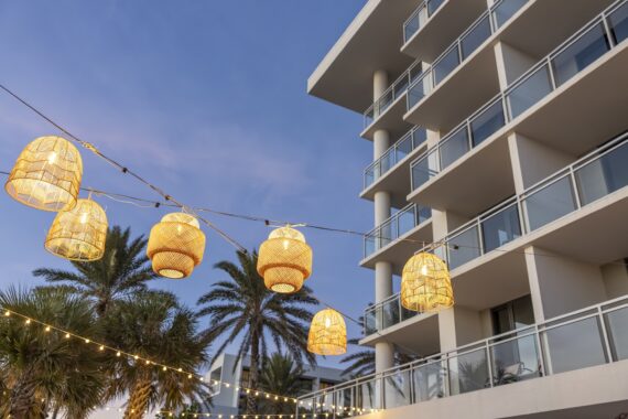 String lights with woven lanterns hang in front of a modern multi-story building with glass balconies, palm trees, and a twilight sky in the background.