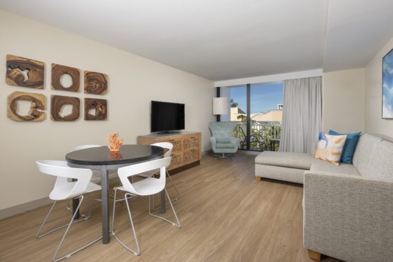 Modern living room with a round dining table and four white chairs, a light gray sectional sofa, a TV on a wooden cabinet, abstract wall art, large windows with curtains, and a blue accent chair by the balcony.