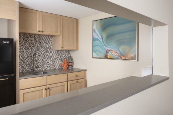 A small kitchen with light wood cabinets, a gray countertop, mosaic tile backsplash, a black refrigerator, a sink, and a colorful abstract painting on the wall. There is a pass-through counter in the foreground.