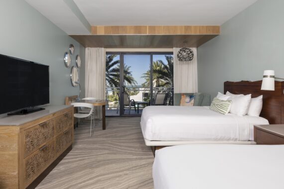 A modern hotel room with two white beds, a wooden dresser with a large TV, a small desk and chair, and a balcony overlooking palm trees and a blue sky. The decor features light colors and beach-inspired details.