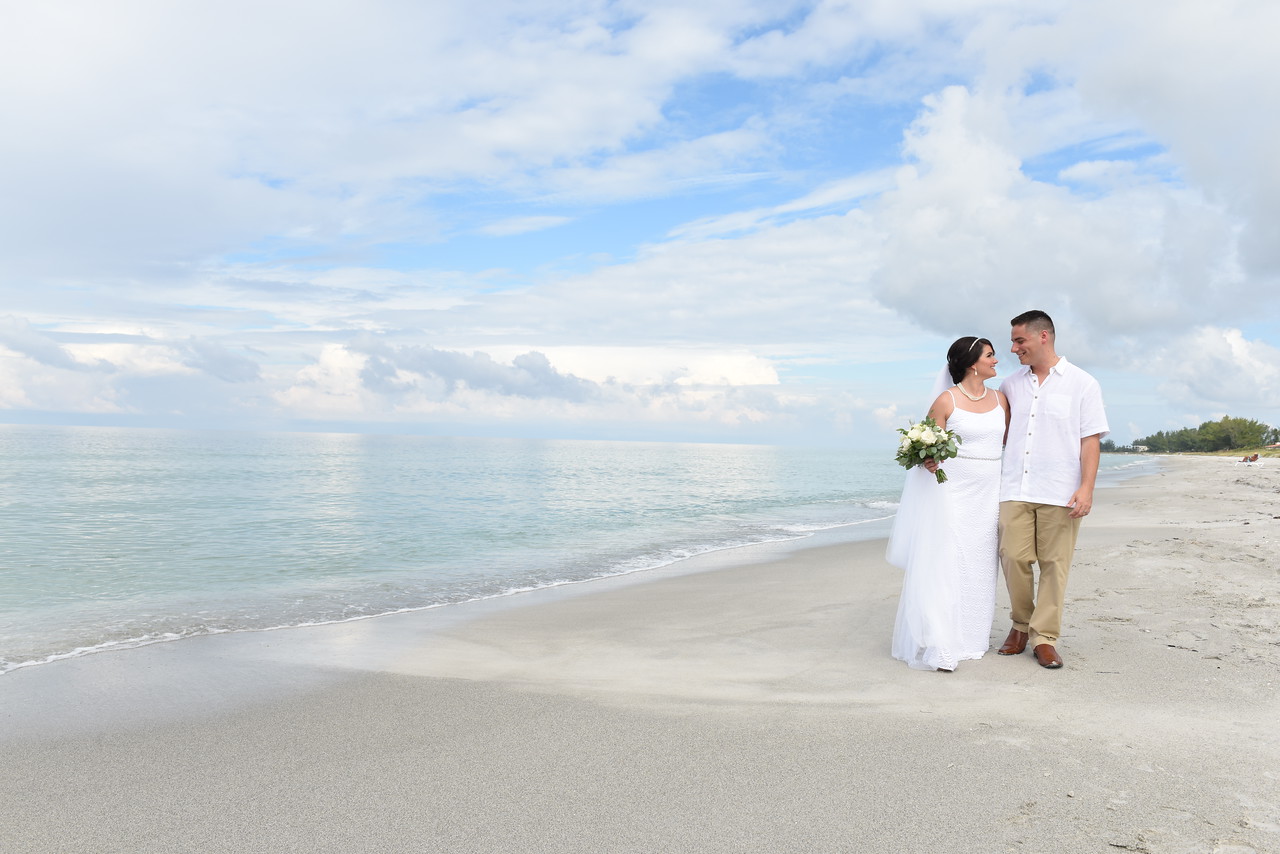 A couple in wedding attire walks hand in hand along a serene beach. The bride holds a bouquet, and both smile at each other with the ocean and cloudy blue sky in the background.
