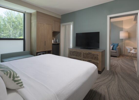 Modern hotel room with a neatly made bed, patterned carpet, large TV on a carved wooden dresser, mini-fridge, closet, sofa by the window, and a view into a separate sitting area with a desk and lamp.