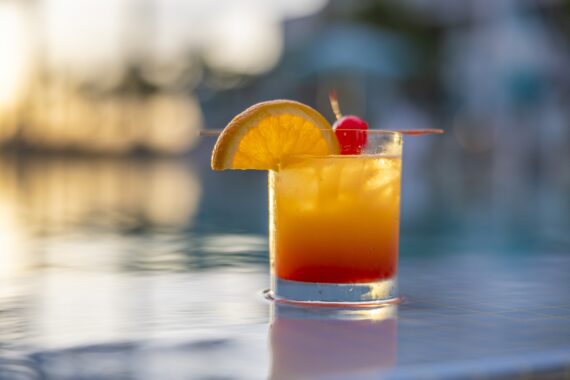 A glass of orange and red cocktail garnished with an orange slice and cherry sits on the edge of a pool, with a blurred background of water and palm trees at sunset.