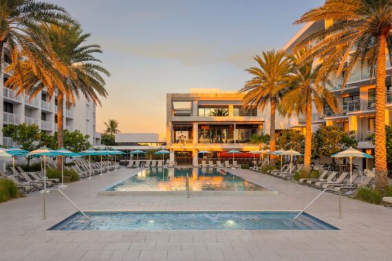 A modern resort pool area at sunset, featuring palm trees, lounge chairs, and umbrellas lining two rectangular swimming pools in front of a contemporary hotel building.
