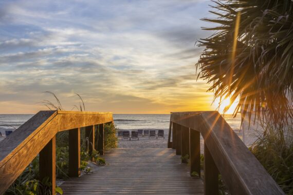 A wooden boardwalk leads to a sandy beach at sunset, with empty lounge chairs facing the ocean and palm leaves framing the scene. Sunlight filters through the foliage, casting a warm glow.