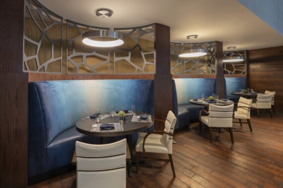 Modern restaurant interior with curved blue upholstered booths, white chairs, round tables set for dining, wooden floors, and decorative wall partitions with geometric patterns under circular ceiling lights.