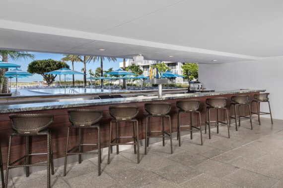 Outdoor poolside bar with wicker stools lined up at a marble counter, facing a view of a pool, palm trees, lounge chairs, and blue umbrellas under a bright sky.