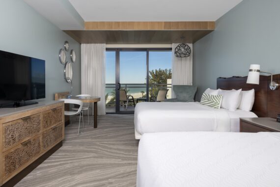 Modern hotel room with two white beds, a large TV on a wooden dresser, a desk and chair by floor-to-ceiling window doors, and a balcony overlooking the ocean and trees. The room has light blue walls and soft, neutral decor.