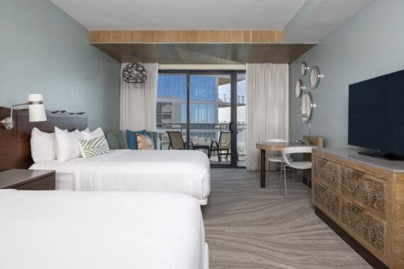 Modern hotel room with two white beds, a large flat-screen TV on a wooden dresser, a small seating area with chairs and a table near a glass balcony door, and neutral decor with light blue walls.