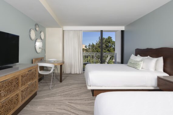 Modern hotel room with two white beds, a wooden dresser with a TV, a desk with a chair, decorative wall mirrors, and a balcony with sliding glass doors and a view of trees outside.