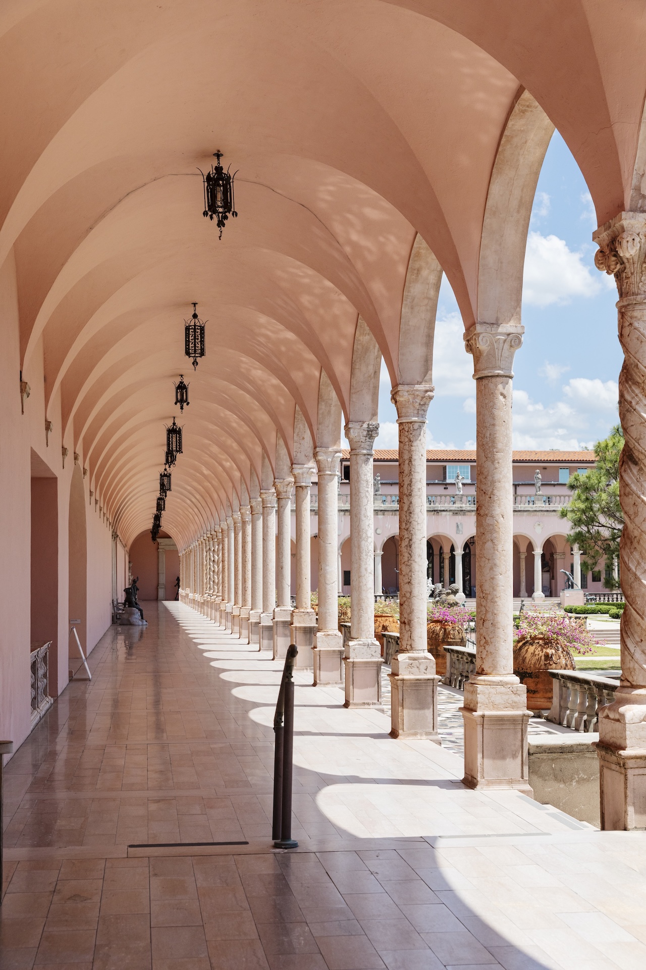 A sunlit arched walkway with pink columns and ornate hanging lamps, overlooking a courtyard garden with trees and buildings under a blue sky.