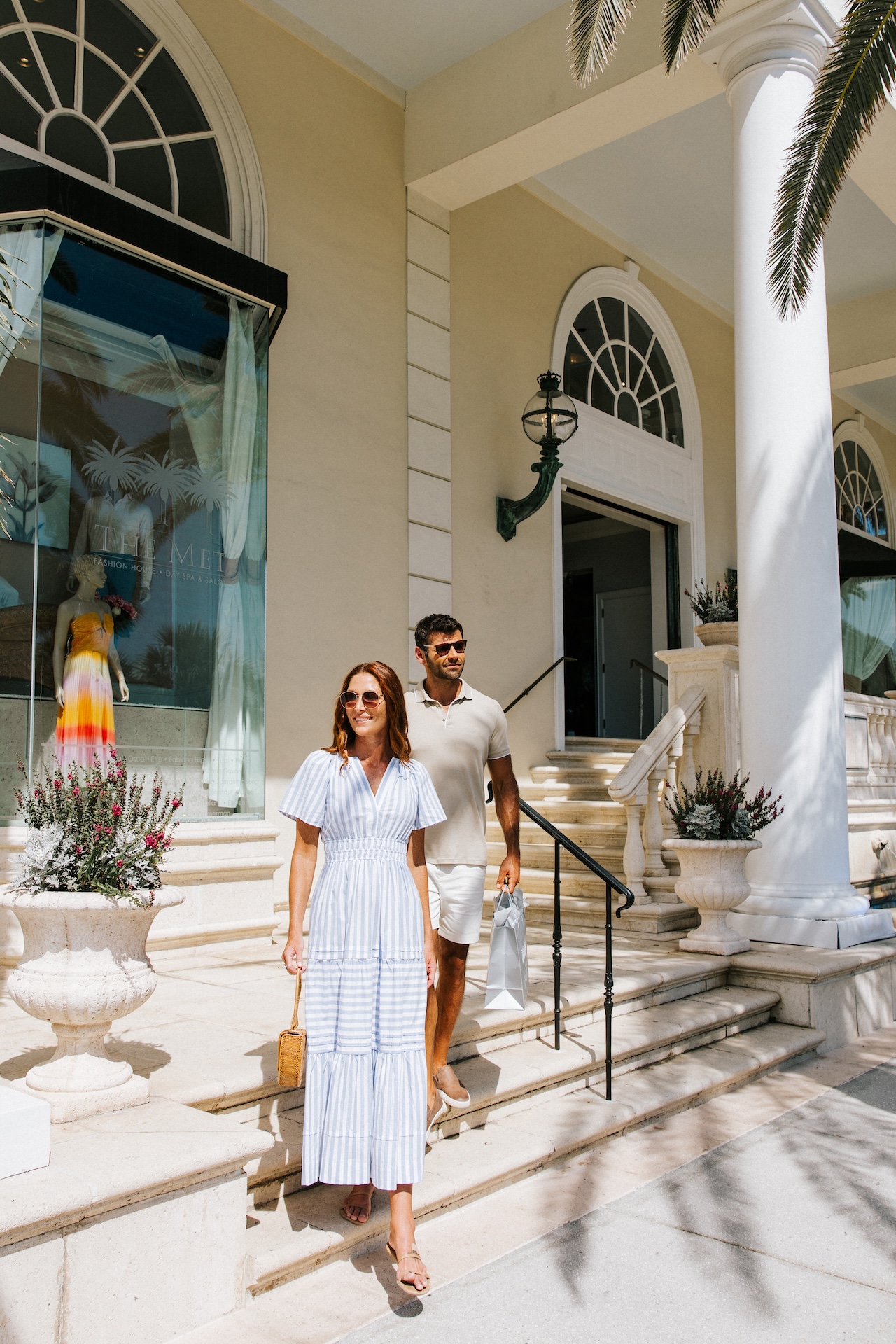 A woman in a white dress and sunglasses walks down steps beside a man in a light outfit, outside a building with arched windows, columns, and a shop displaying a colorful dress.