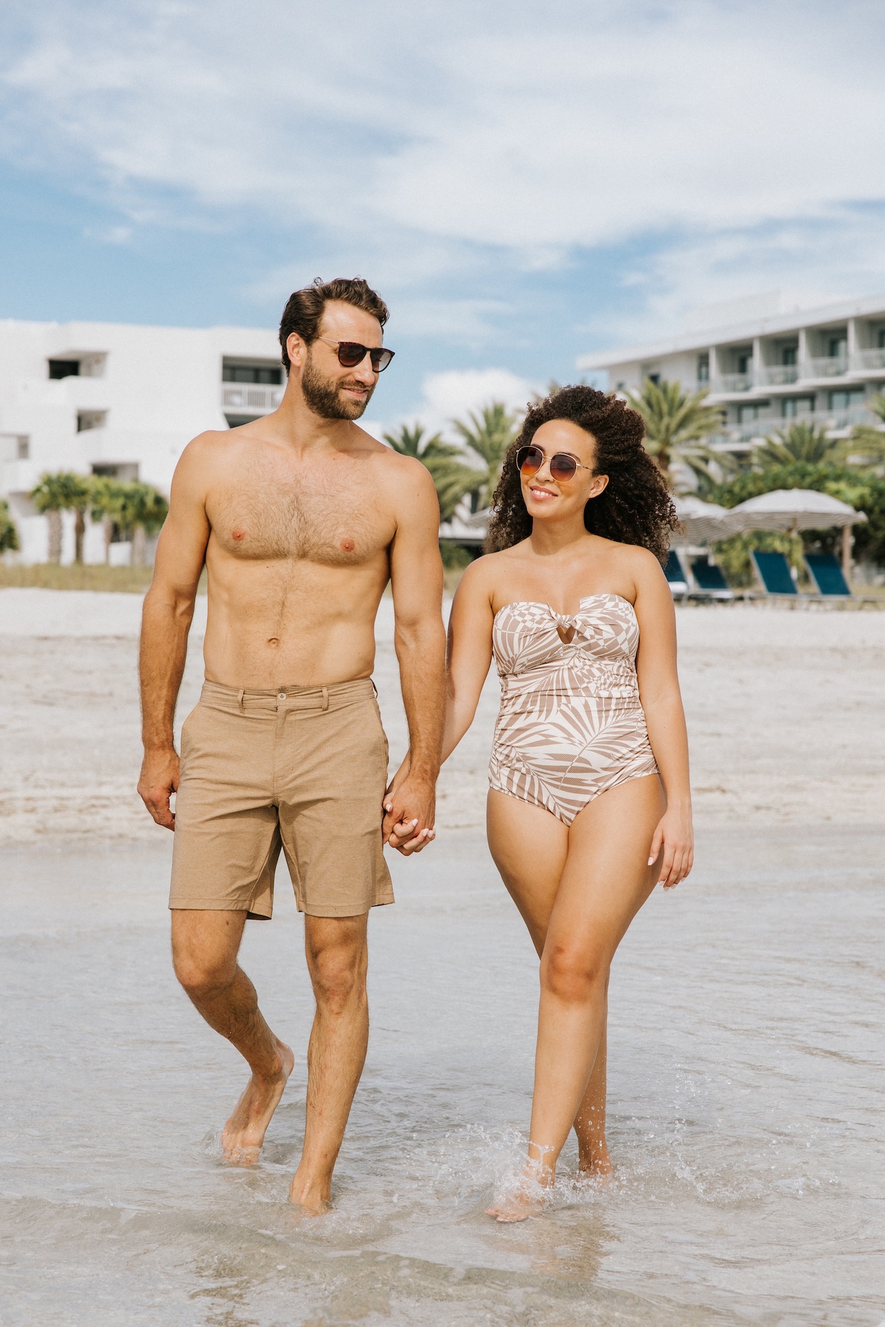 A man and woman in swimwear hold hands while walking at the beach. They are smiling, wearing sunglasses, and there are palm trees, lounge chairs, and buildings in the background.