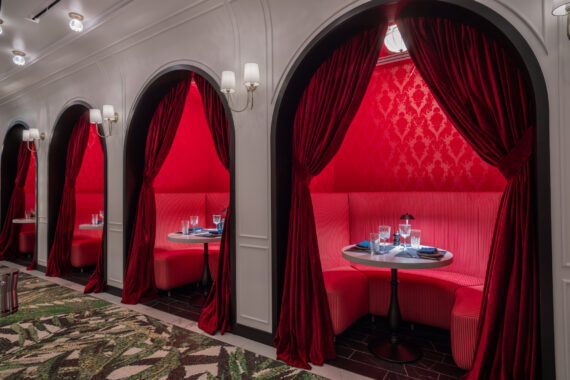 Three elegant red velvet booths with round tables and place settings are set within arched alcoves. Rich curtains frame each booth, creating a cozy, luxurious dining atmosphere. Patterned carpet runs along the floor.