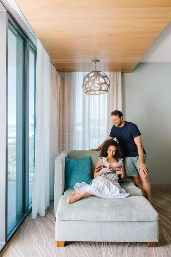 A woman sitting on a chaise lounge reading a magazine while a man stands beside her, both smiling in a modern hotel room.