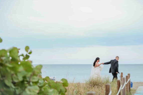 A couple walks hand in hand on a sandy beach path. The bride is in a white dress and the groom in a black suit. Green foliage and dune grass surround the path, with the ocean and a cloudy sky in the background.