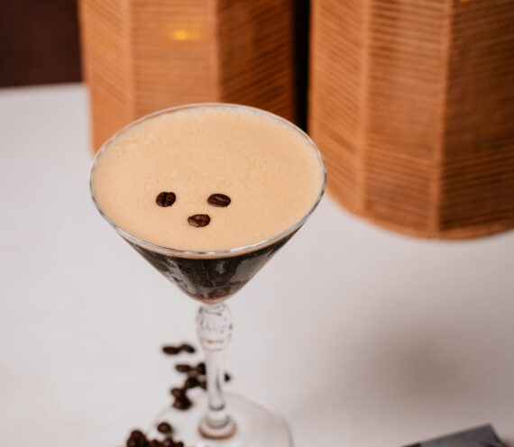 A martini glass filled with a foamy espresso martini, garnished with three coffee beans, sits on a white surface next to scattered coffee beans and a black-and-white magazine. Two wicker lanterns are in the background.