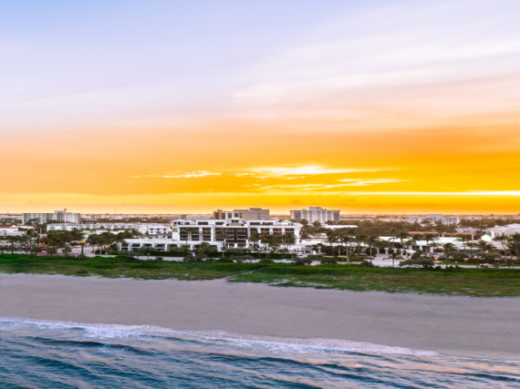 Aerial view of a beach with gentle waves in the foreground, a line of green vegetation, and buildings in the midground. The sky is filled with a vibrant orange and yellow sunset.