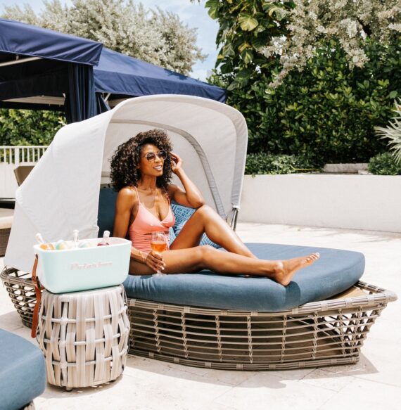 A woman in a pink swimsuit and sunglasses sits on a cushioned outdoor lounge chair with a canopy, holding a drink. A cooler filled with items is beside her, and there are trees and a patio in the background.