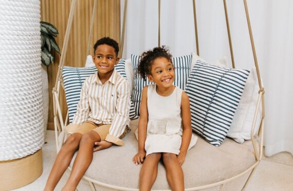 Two young children sit and smile on a round hanging chair with striped cushions. The boy wears a striped shirt and tan shorts; the girl wears a light-colored sleeveless dress. The setting is bright and cozy with neutral decor.