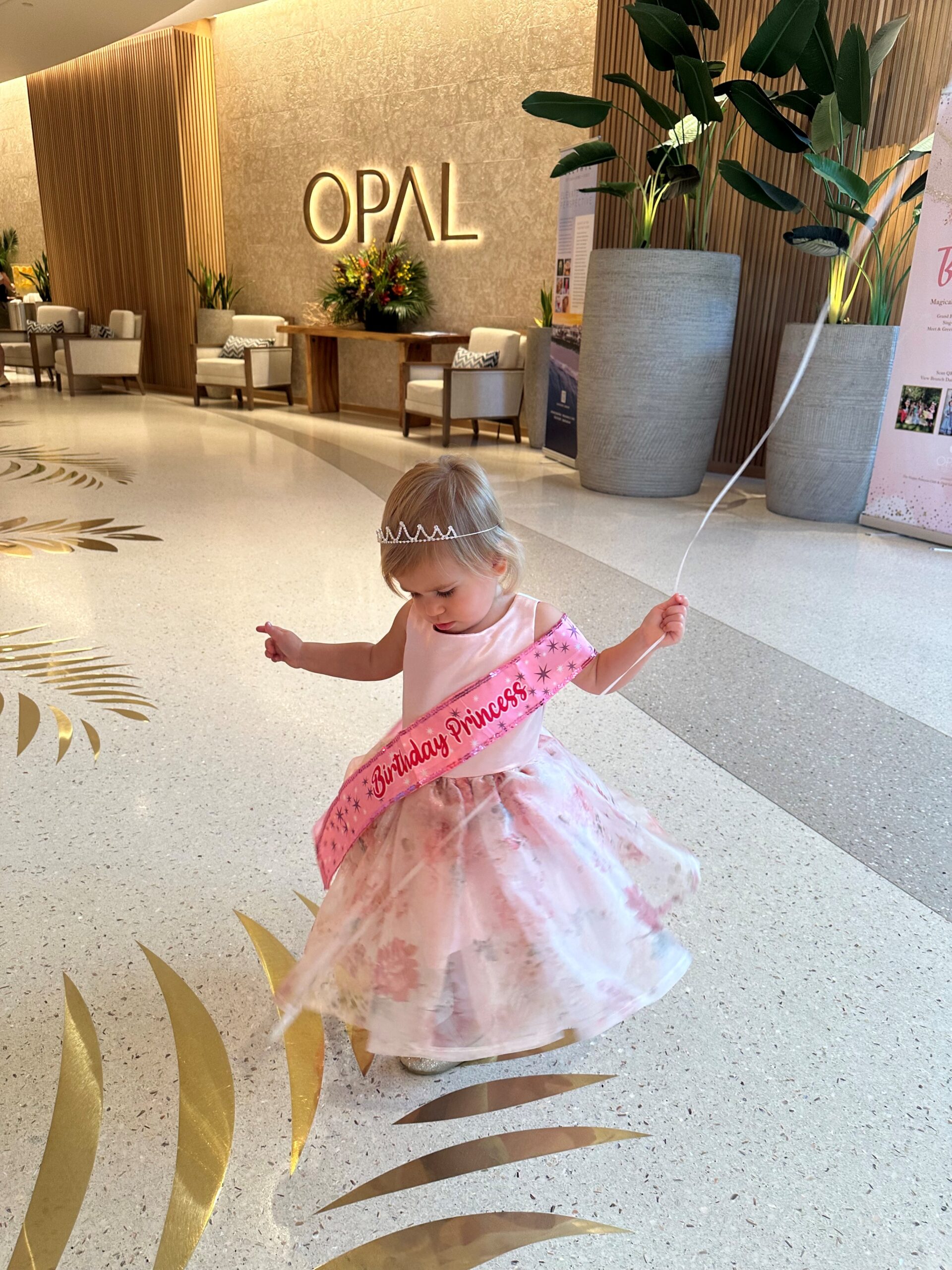 A young girl wearing a pink floral dress, tiara, and Birthday Princess sash twirls with a balloon in a bright, elegant lobby with large plants and OPAL signage on the wall.