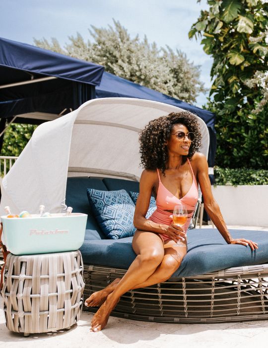 A woman in a pink swimsuit sits on a blue outdoor lounge chair, smiling and holding a drink. There’s a cooler with drinks beside her, and greenery and a sunshade are in the background.